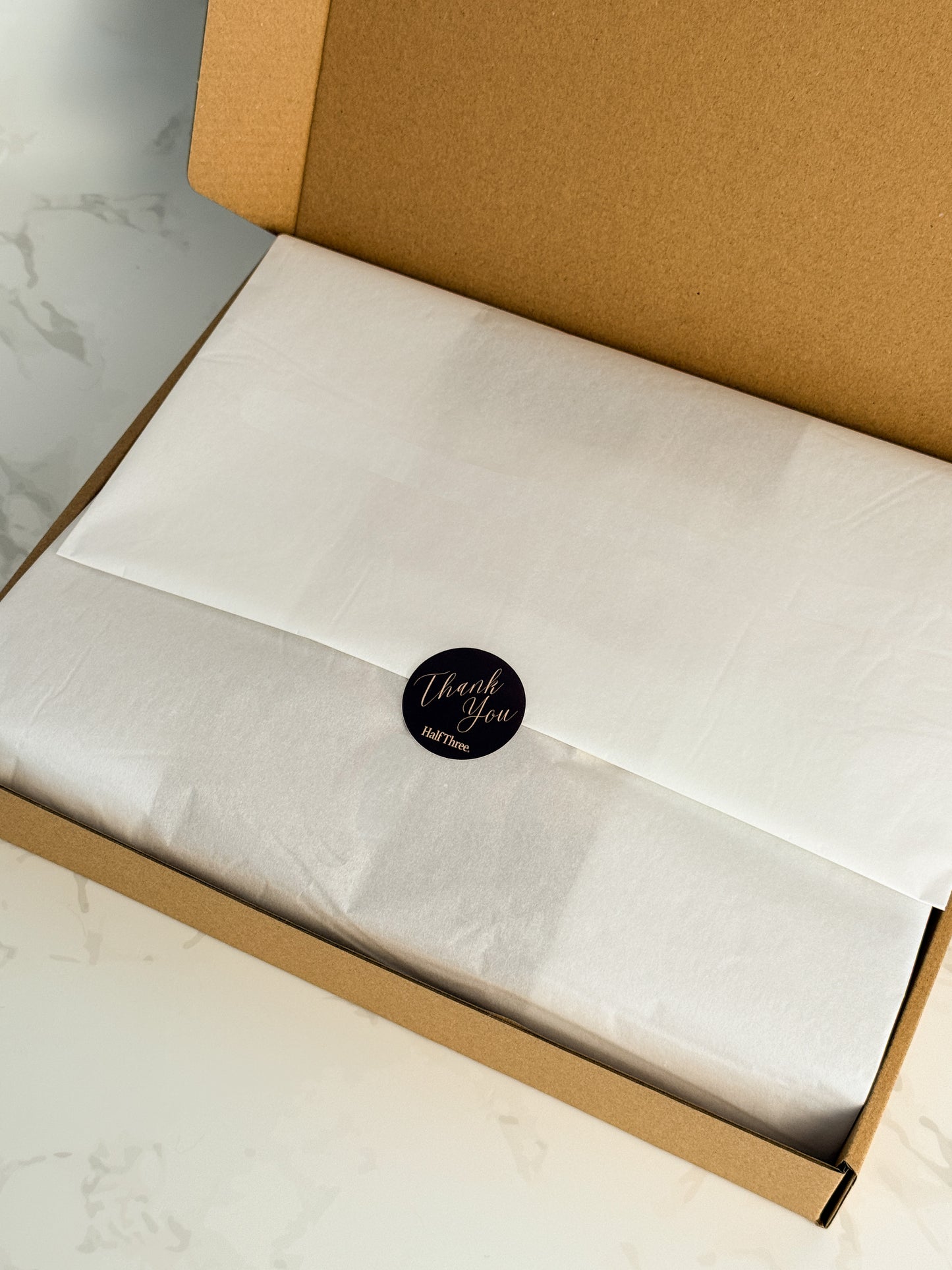 An open brown cardboard gift box containing white tissue paper and a small round brown 'Thank You' sticker with the Half Three logo.