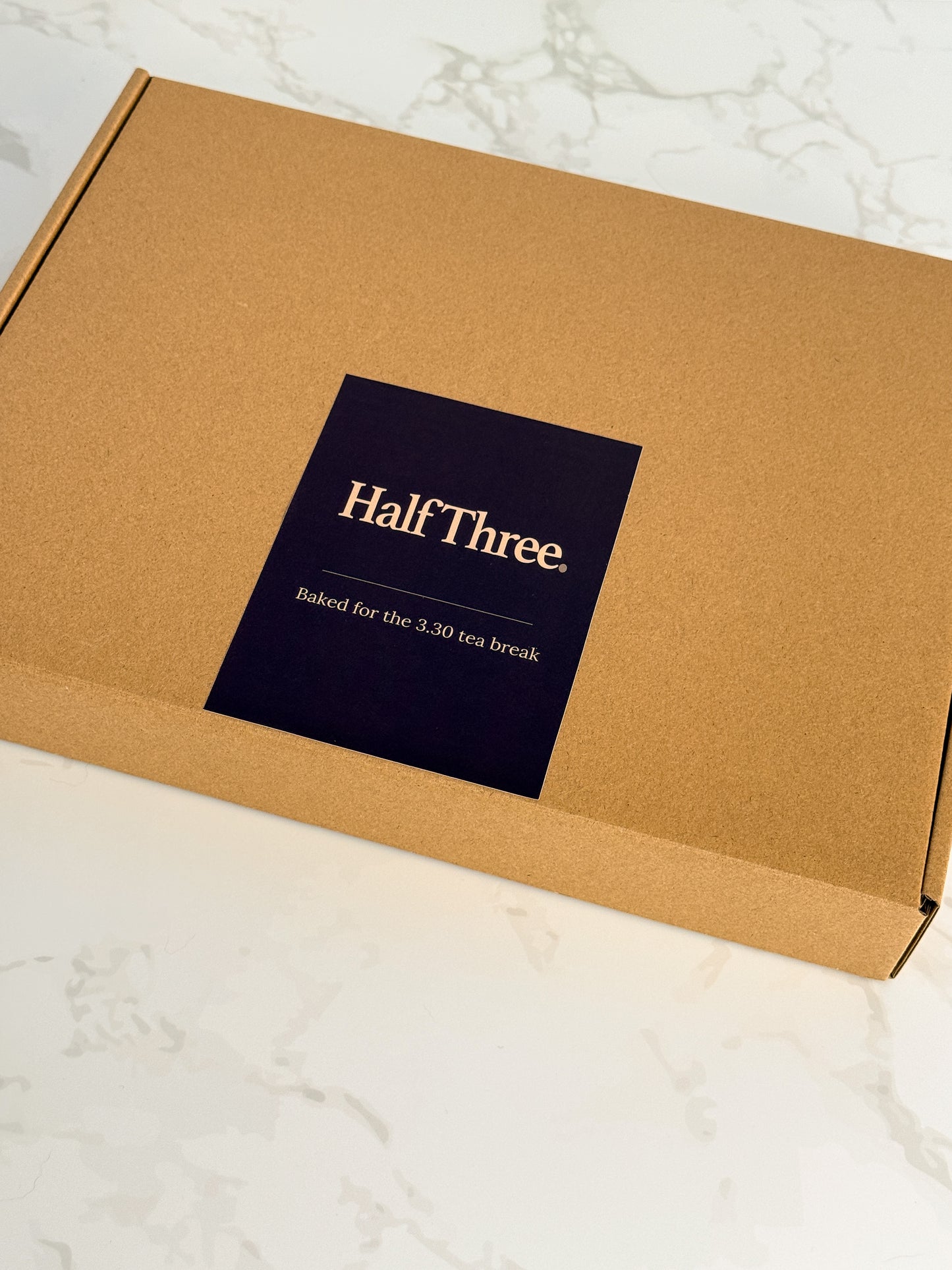 A square kraft paper box with a dark navy label reading 'Half Three. Baked for the 3:30 tea break' in gold text, photographed on a light marble surface.