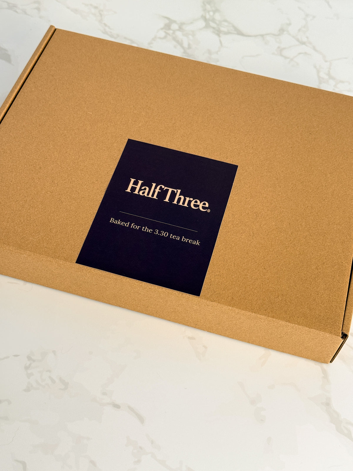 A square kraft paper box with a dark navy label reading 'Half Three. Baked for the 3:30 tea break' in gold text, photographed on a light marble surface.
